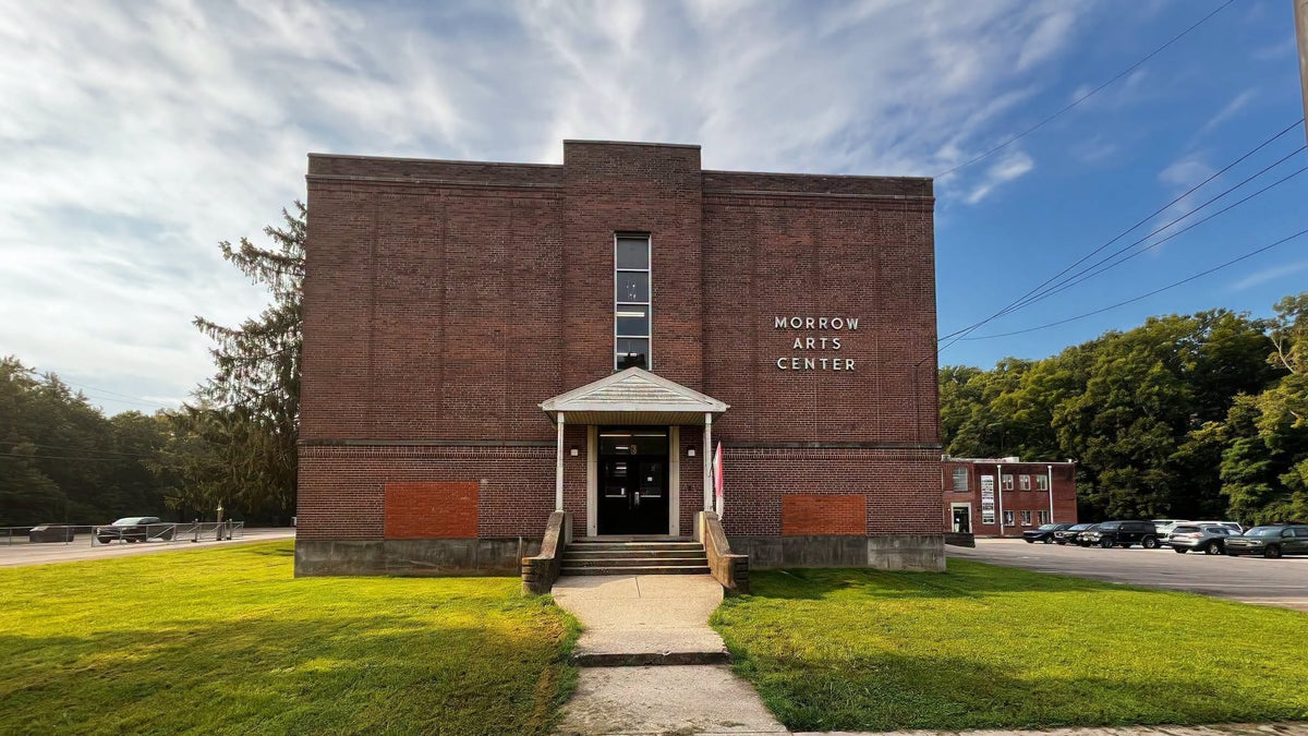 The front exterior of the Morrow Arts Center brick building, featuring a central entrance with steps, a concrete walkway leading up the path, green grass, trees, and a blue sky with clouds.